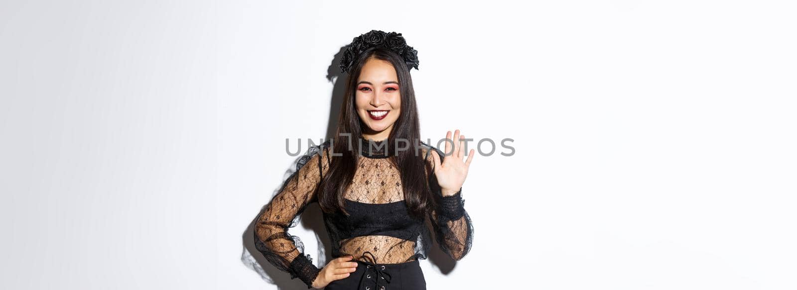 Image of friendly stylish asian woman in gothic lace dress waving hand to say hello, greeting someone, welcome people at halloween party, standing over white background by Benzoix