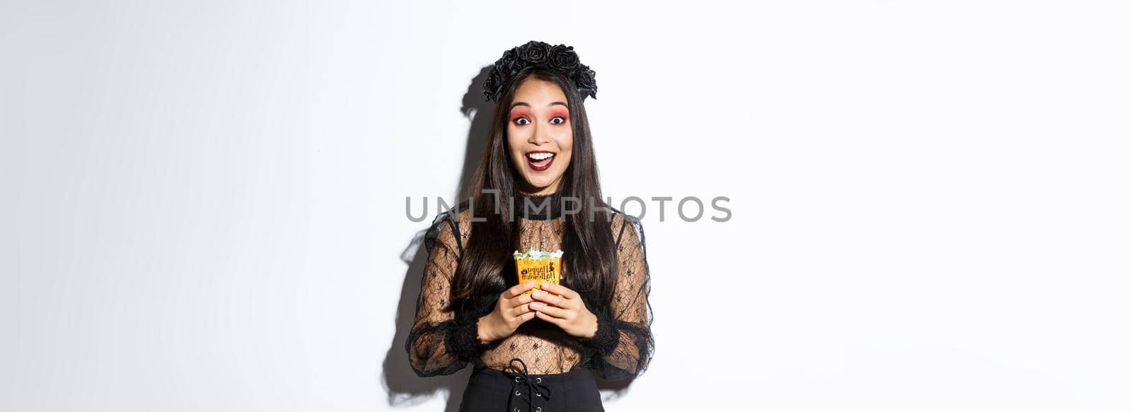 Beautiful asian girl smiling happy, holding sweets, wearing witch costume on halloween, enjoying trick or treating.