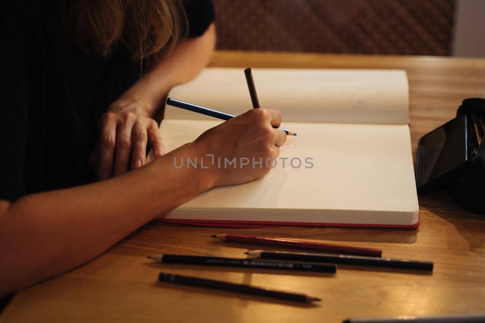 A girl in a cafe draws in a sketchbook with colored pencils. Notepad, pencils, black pencil case.