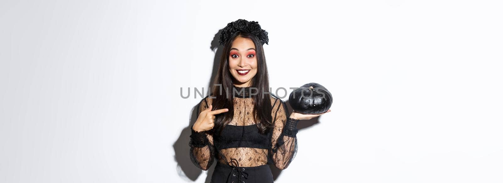 Surprised happy asian woman in witch costume pointing finger at black pumpkin, smiling amused, getting ready for halloween party, standing over white background.