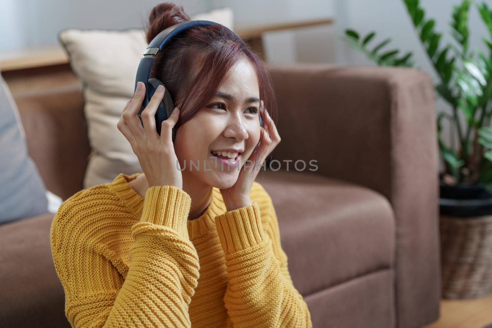 A portrait of a young Asian woman with a smiling face wearing a pair of headphones and using computer and listening to music while sitting on the sofa by Manastrong