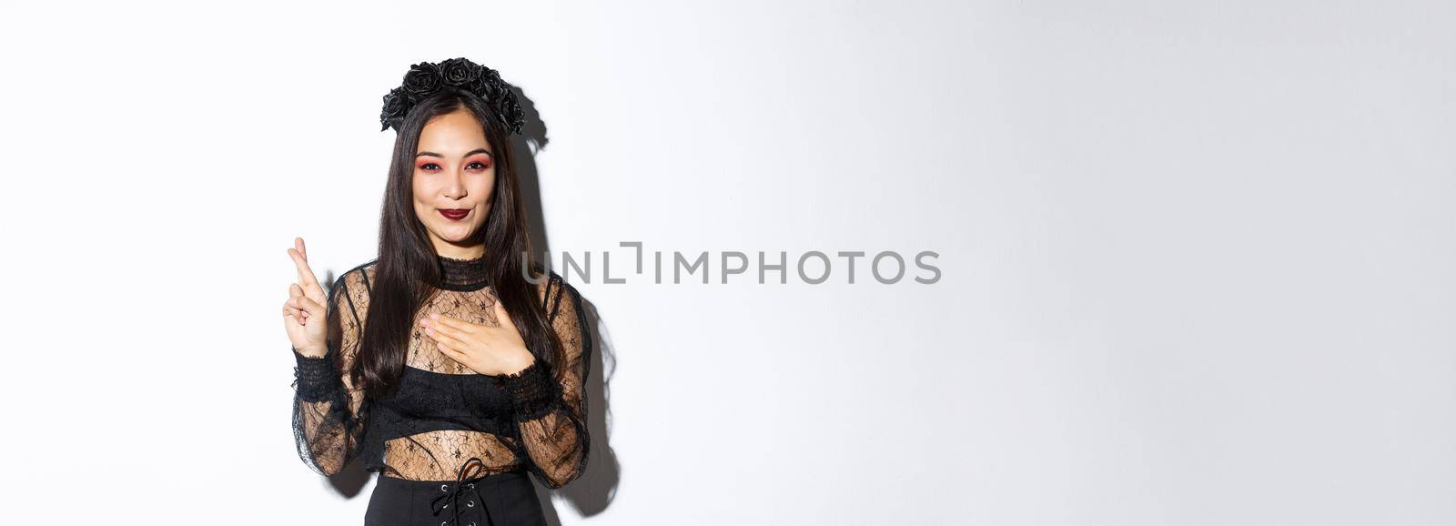 Image of attractive asian woman in halloween party dress making wish, holding hand on heart and cross fingers for good luck, standing over white background.