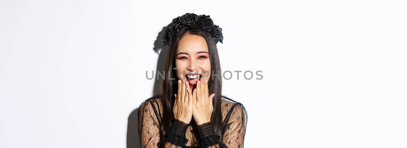 Close-up of happy asian woman celebrating halloween in witch costume and laughing, standing over white background by Benzoix