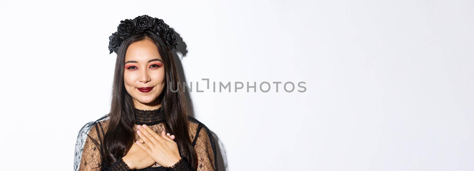 Close-up of thankful smiling asian woman looking grateful with hands over chest, standing in witch costume over white background.