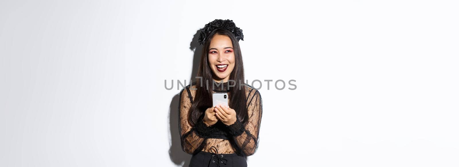 Happy attractive asian woman in halloween costume laughing carefree, holding smartphone and looking amused at upper left corner, standing over white background.