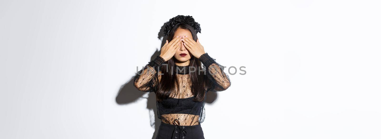 Image of woman wearing gothic wreath and black lace dress shut her eyes with hands, waiting for surprise on halloween, standing over white background with patient expression.