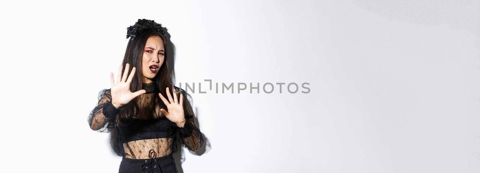 Image of bothered and annoyed asian woman in elegant gothic dress raising hands defensive, grimacing from camera flesh, asking to stop taking pictures of her, standing white background.