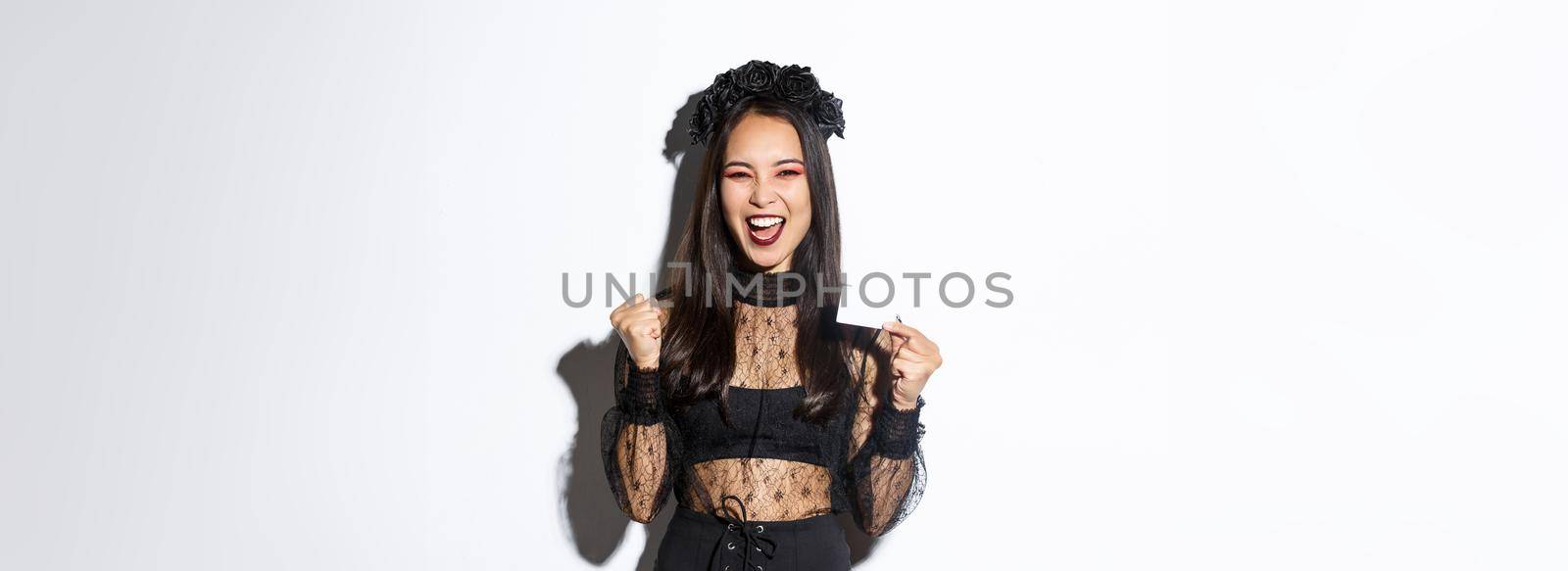 Cheerful asian woman in gothic lace dress and black wreath showing credit card and rejoicing, standing over white background by Benzoix