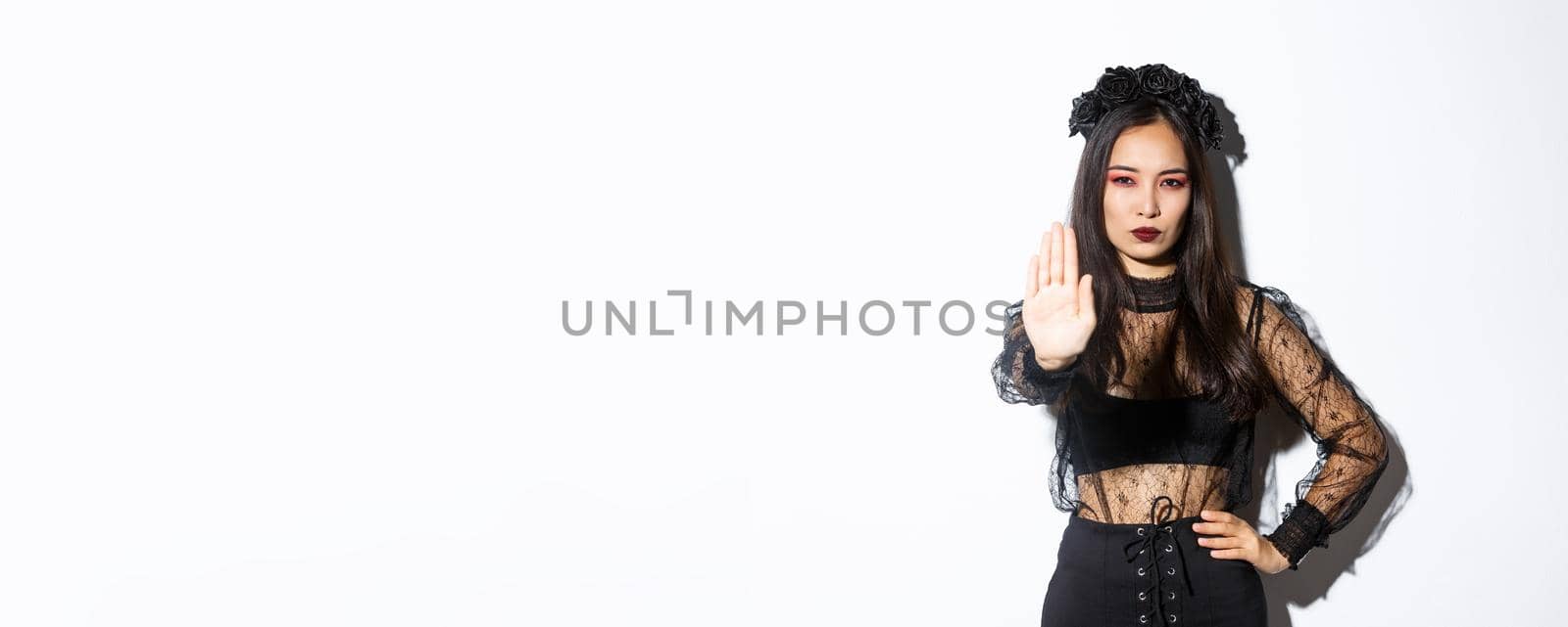 Image of serious asian woman in halloween costume of witch, showing stop gesture, forbid or prohibit something with displeased confident face, standing white background.