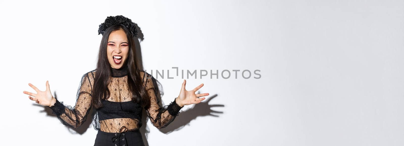 Woman screaming and looking scary in her halloween witch costume, spread hands sideways and looking angry, trying scare people during trick or treat, standing over white background.