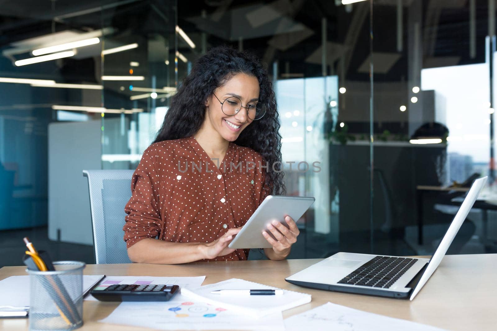 Young successful business woman working inside modern office building, Hispanic woman using tablet computer smiling and happy.