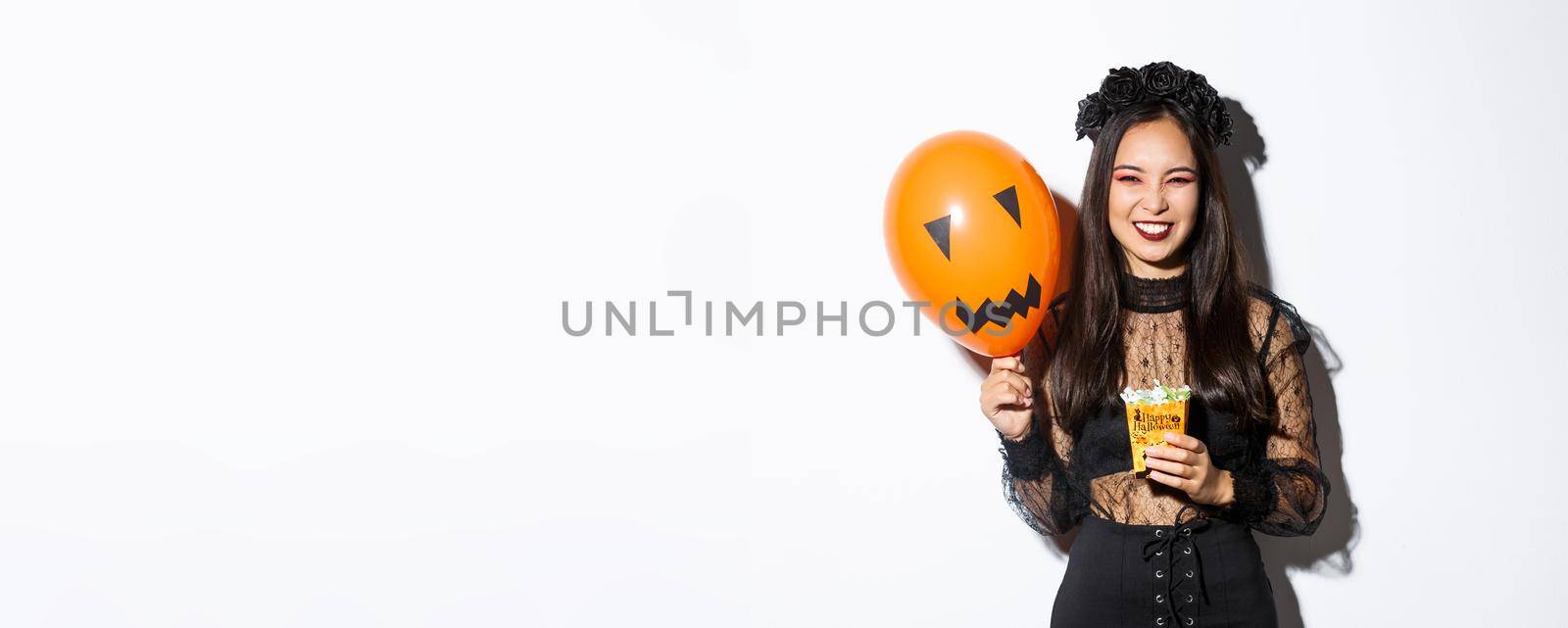 Sassy beautiful asian woman enjoying trick or treating, celebrating halloween, holding orange balloon and sweets by Benzoix