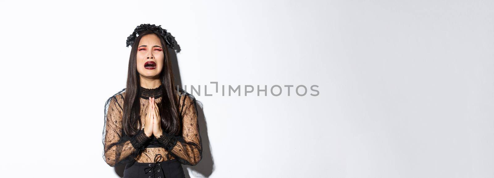 Miserable young asian woman in trouble pleading god, crying and begging for help, wearing halloween gothic dress and wreath, supplicating over white background.