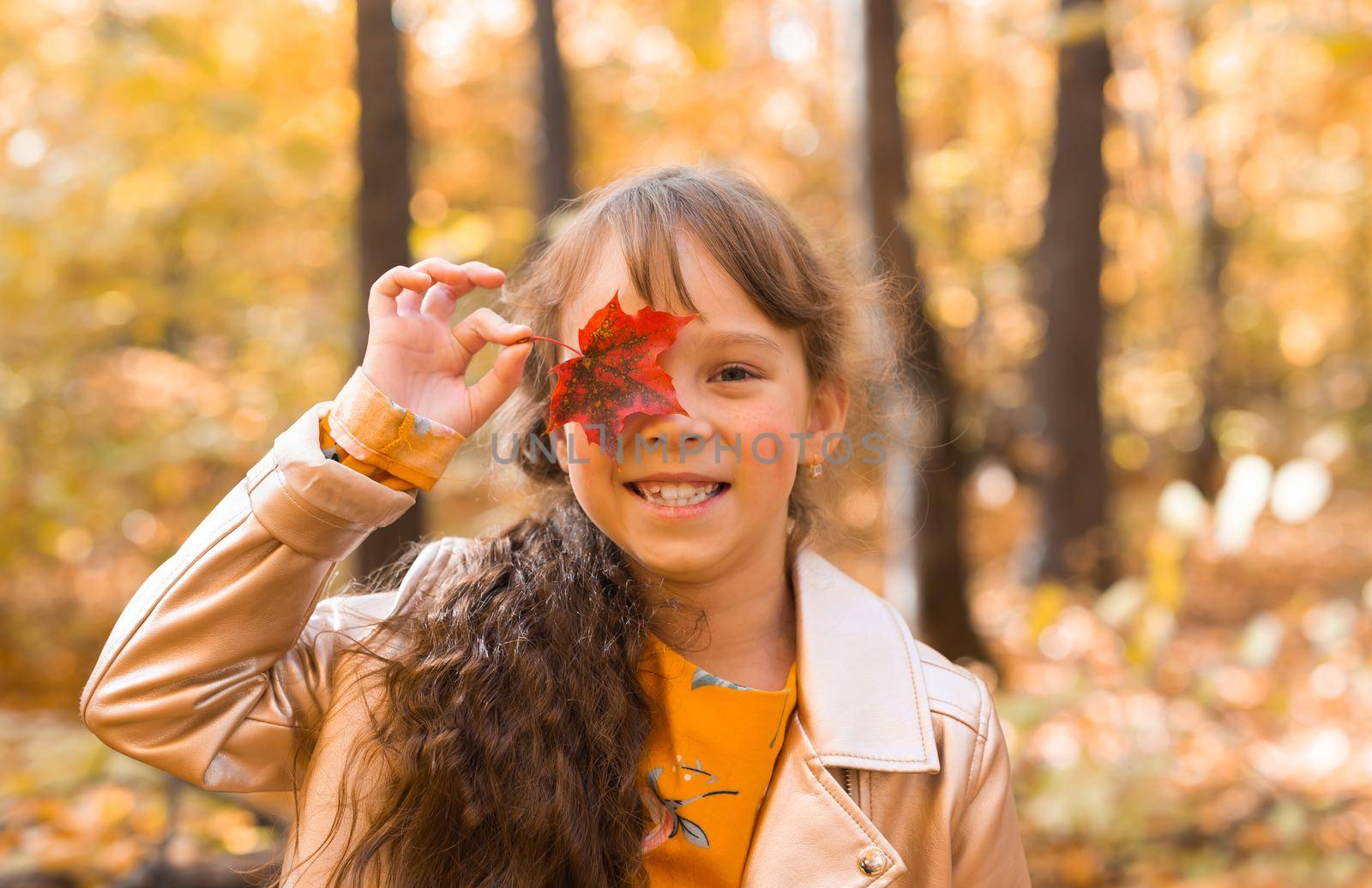 Close portrait of a little girl. She covers her eye with a maple autumn leaf. Fall season and children concept