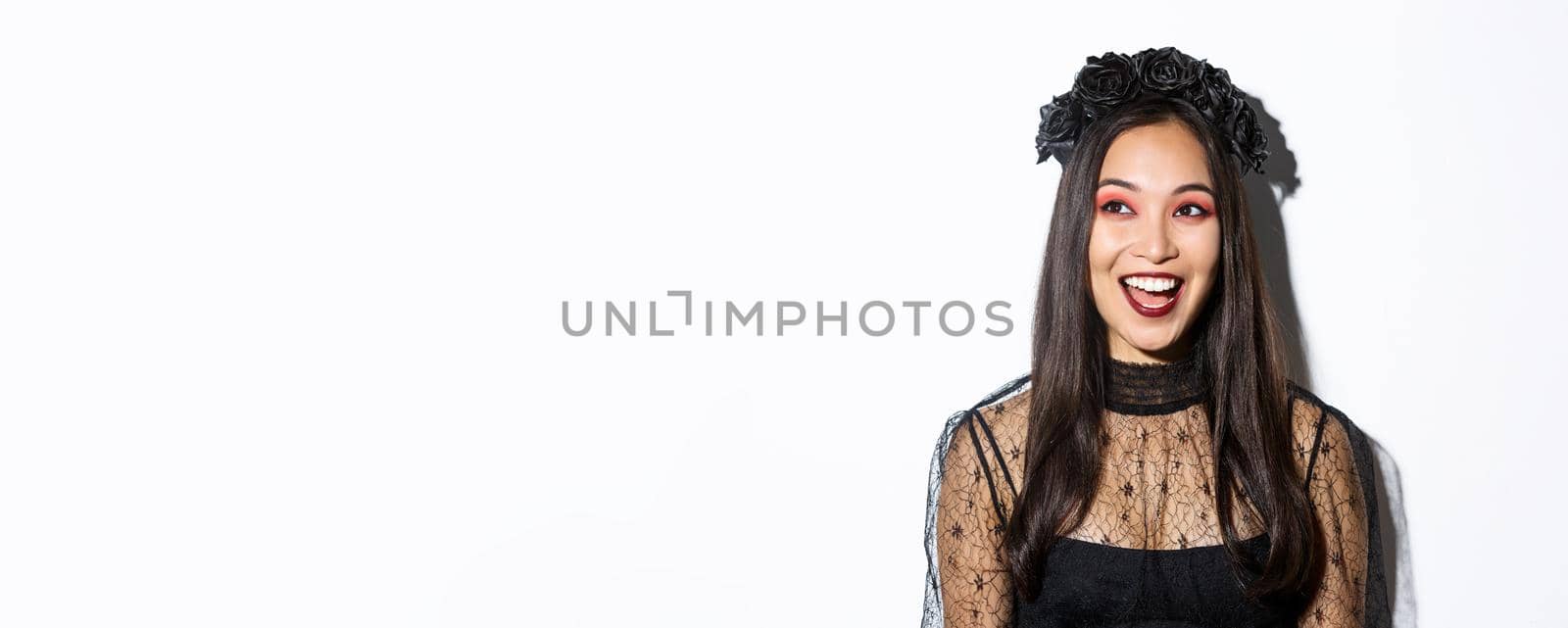 Close-up of happy pretty asian woman in halloween costume looking excited at upper left corner, standing over white background.