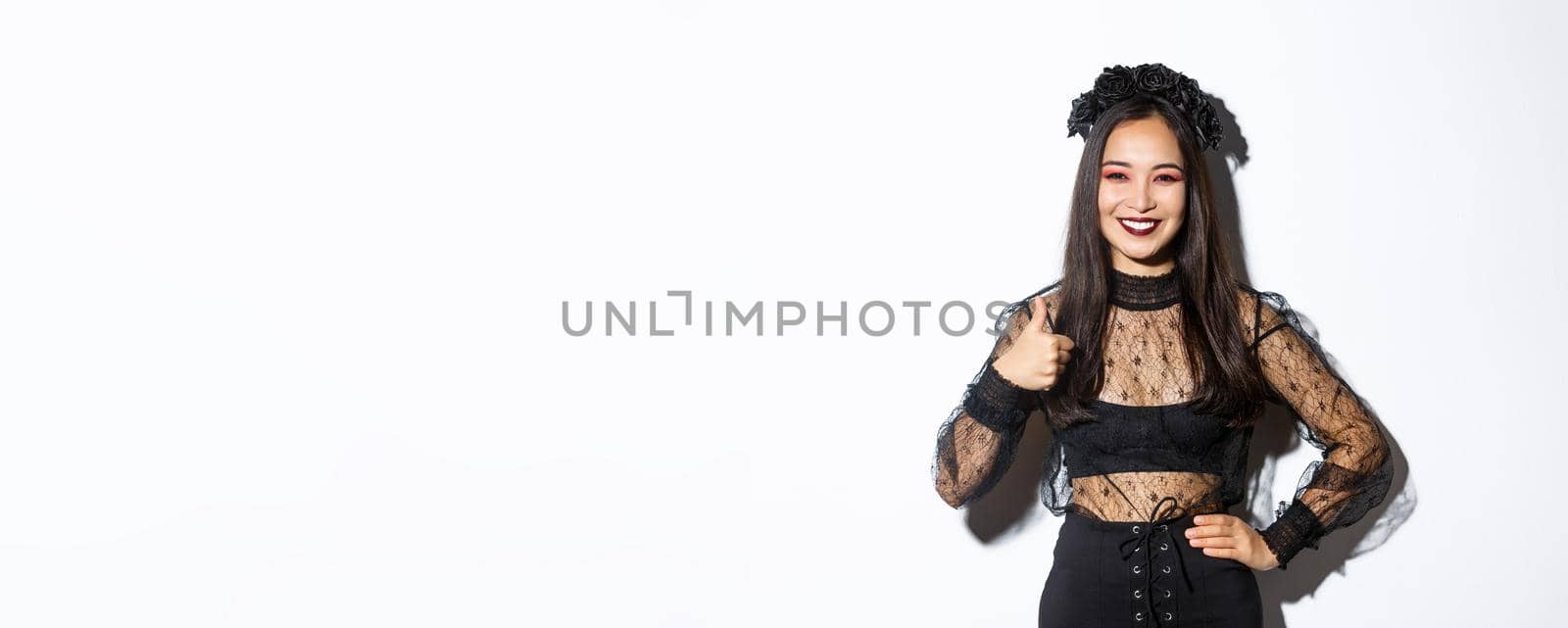 Image of satisfied pretty asian girl in halloween costume showing thumbs-up in approval, smiling pleased. Woman in evil witch party dress looking happy, like something, white background by Benzoix