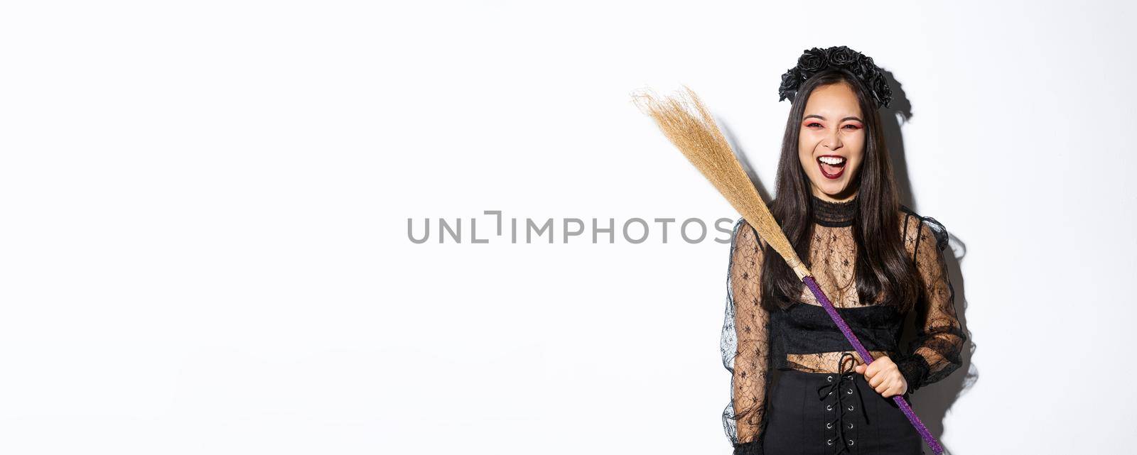Sassy evil witch laughing and waving her broom, wearing halloween costume, standing over white background.