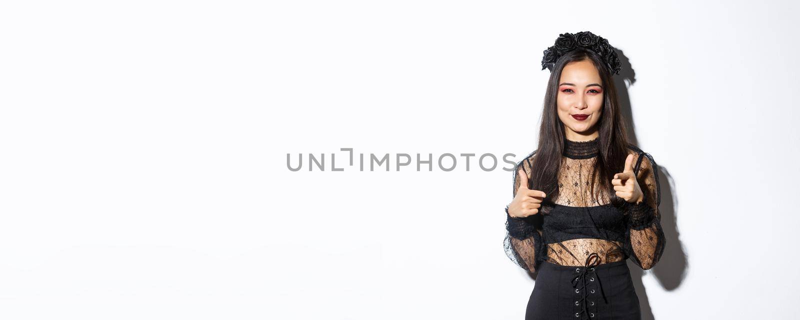 Cheeky and happy attractive woman in witch outfit pointing fingers at camera and smiling, making compliment or praising something good, standing in halloween costume over white background by Benzoix