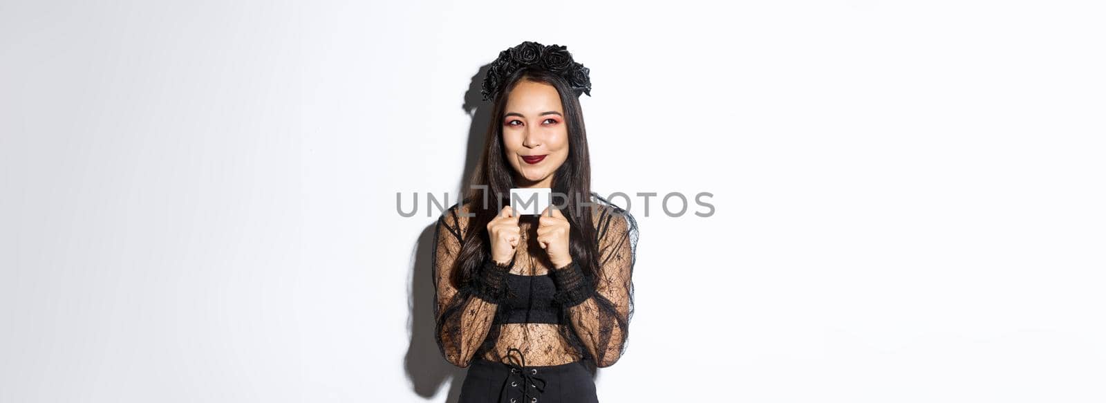 Image of thoughtful asian woman in black gothic dress have a plan, holding credit card, smiling pleased at upper left corner.