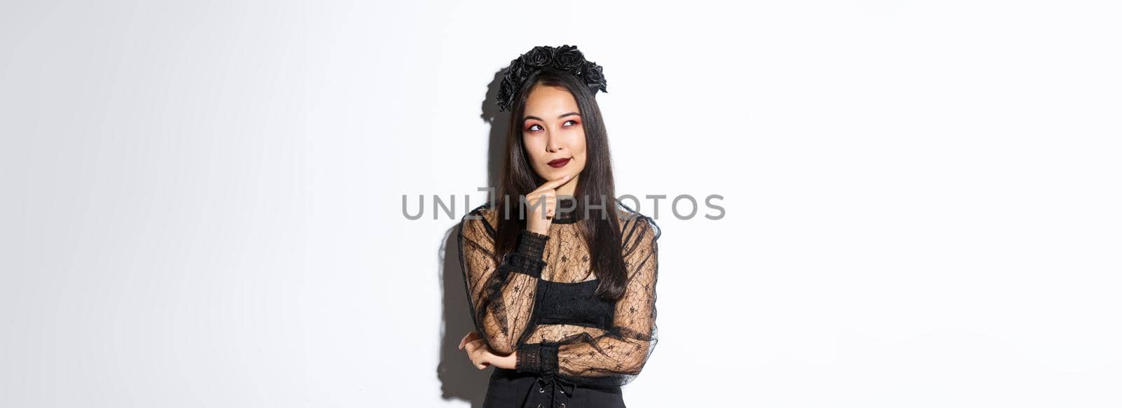 Thoughtful beautiful asian woman standing in black lace dress and gothic wreath, looking upper left corner intrigued. Girl in halloween costume making choice.