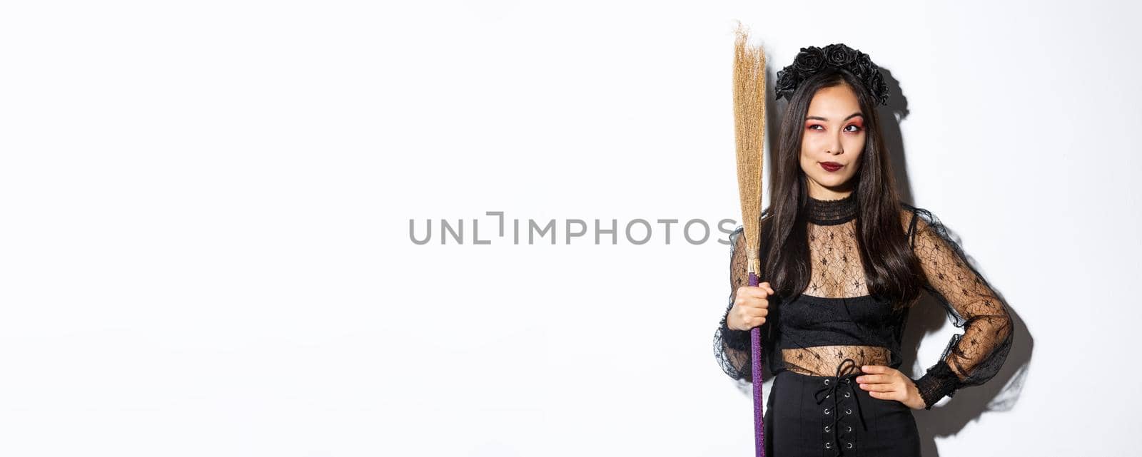 Portrait of smart thoughtful asian girl looking at upper left corner with pleased smirk, holding broom, wearing witch costume for halloween party, standing over white background by Benzoix