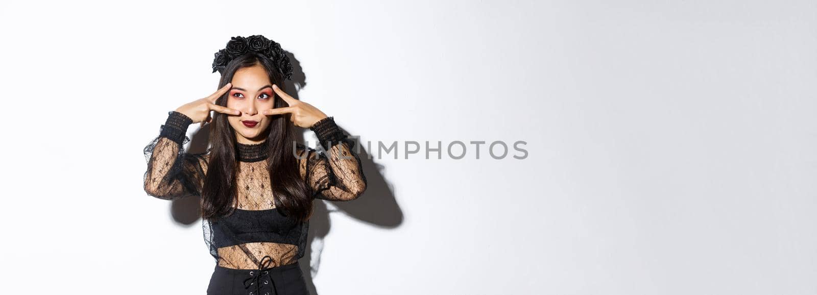Beautiful asian woman in halloween party outfit looking at upper left corner, making peace gestures over eyes, standing in gothic lace dress with black wreath over white background by Benzoix