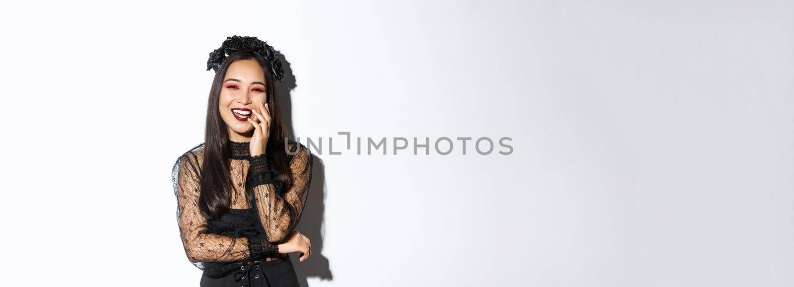 Image of happy good-looking asian woman in gothic dress and black wreath, laughing at halloween party, standing over white background by Benzoix