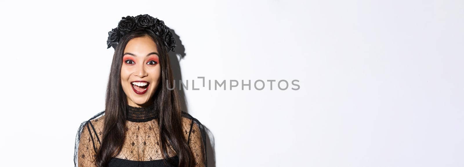 Close-up of enthusiastic asian woman looking amused, wearing witch costume on halloween, standing over white background.