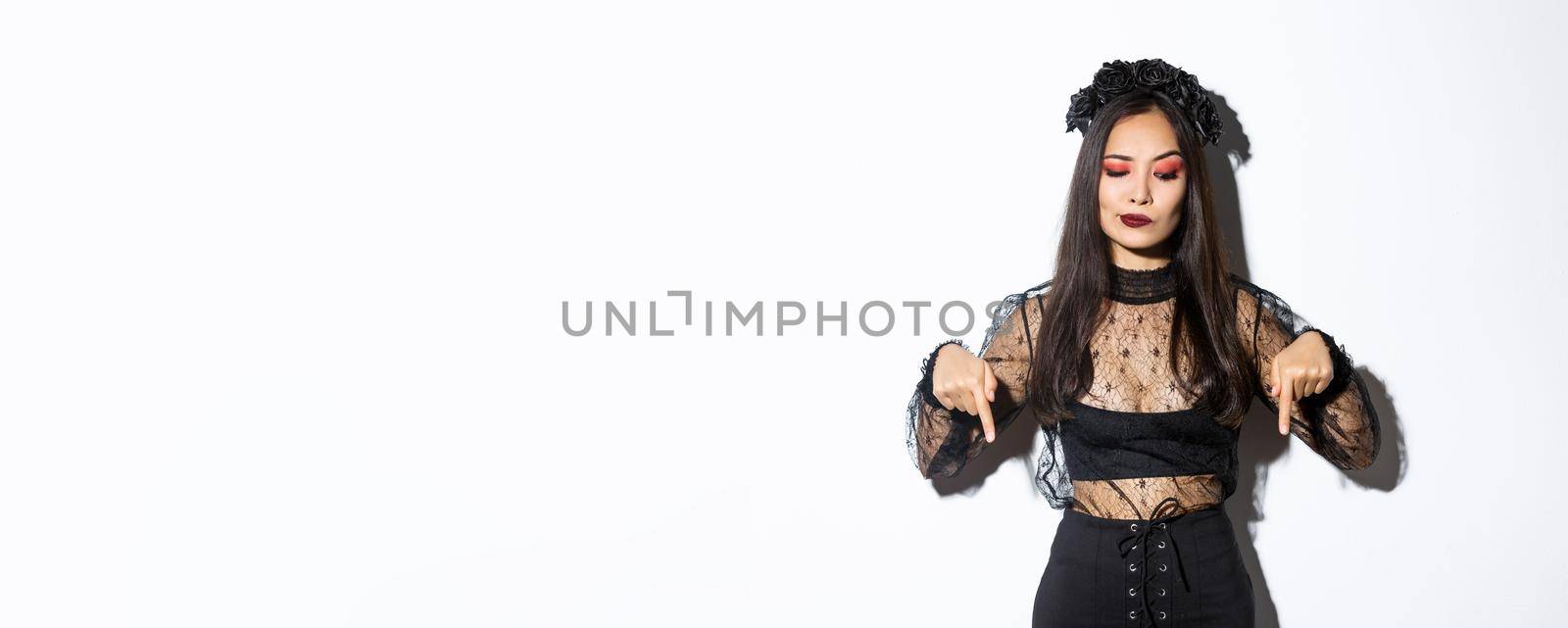 Hesitant and serious-looking asian female in magician costume, dressed-up as evil witch for halloween, looking and pointing fingers down with disbelief, standing over white background by Benzoix