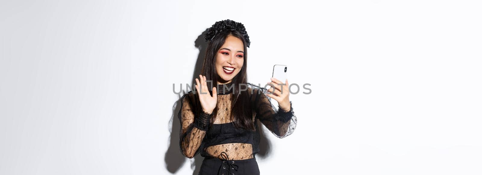Portrait of beautiful and stylish asian woman in gothic lace dress saying hi, waving hand at smartphone camera during video call, standing over white background.