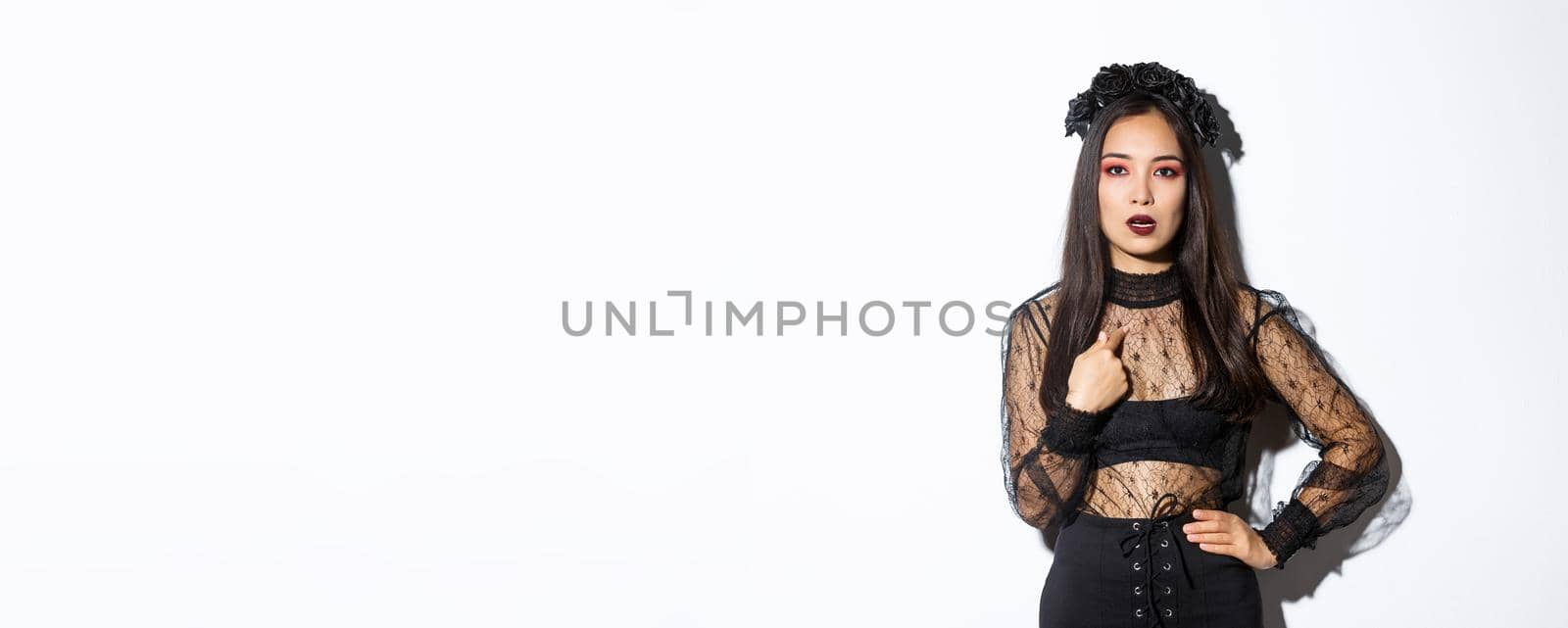 Annoyed and confused asian woman in witch halloween costume pointing at herself, looking bothered and unamused, standing reluctant over white background.