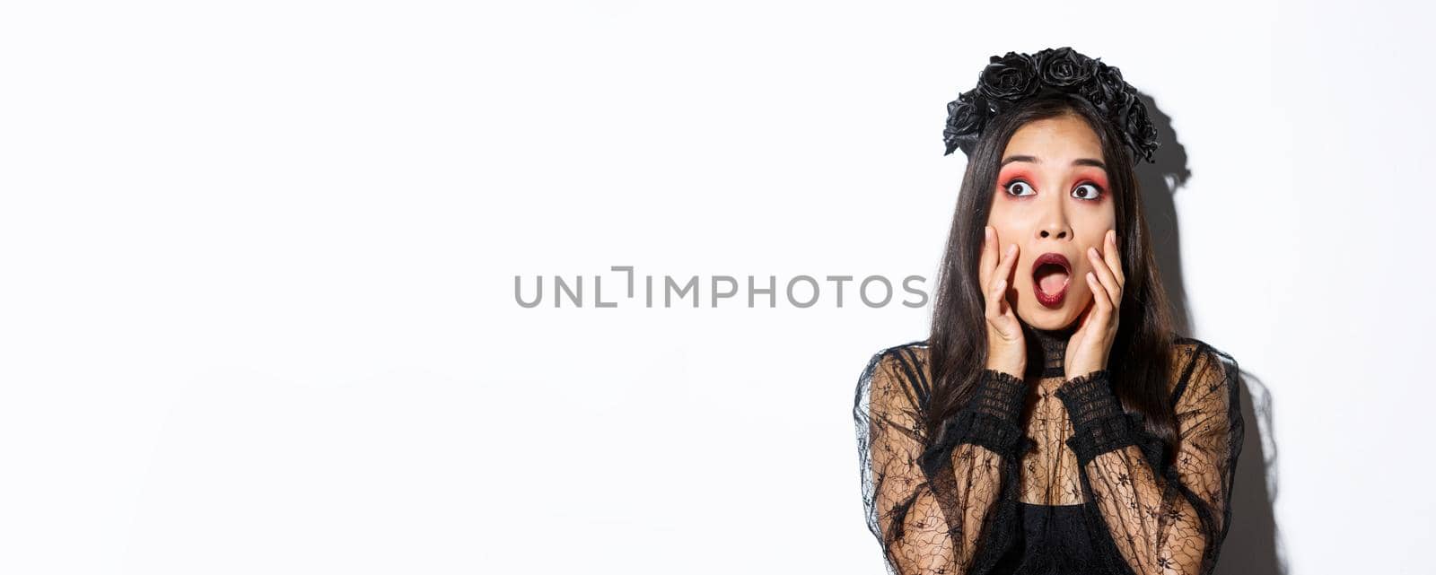 Close-up of scared asian woman in halloween costume of witch looking frightened at upper left corner, gasping and standing ambushed over white background by Benzoix