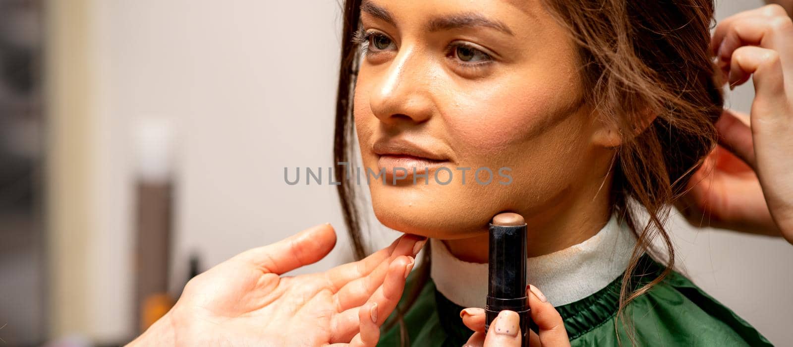 Beautiful young brunette woman receiving makeup with stick concealer on her face in a beauty salon