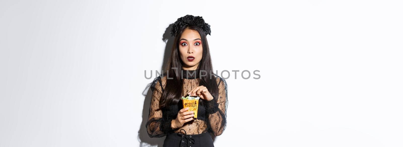 Image of surprised and fascinated asian woman in witch costume, holding sweets gathered during trick or treating, standing over white background.