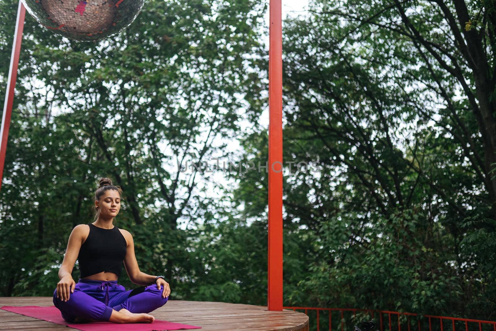 Young woman is practising yoga sitting in the lotus position on a wooden podium in the garden