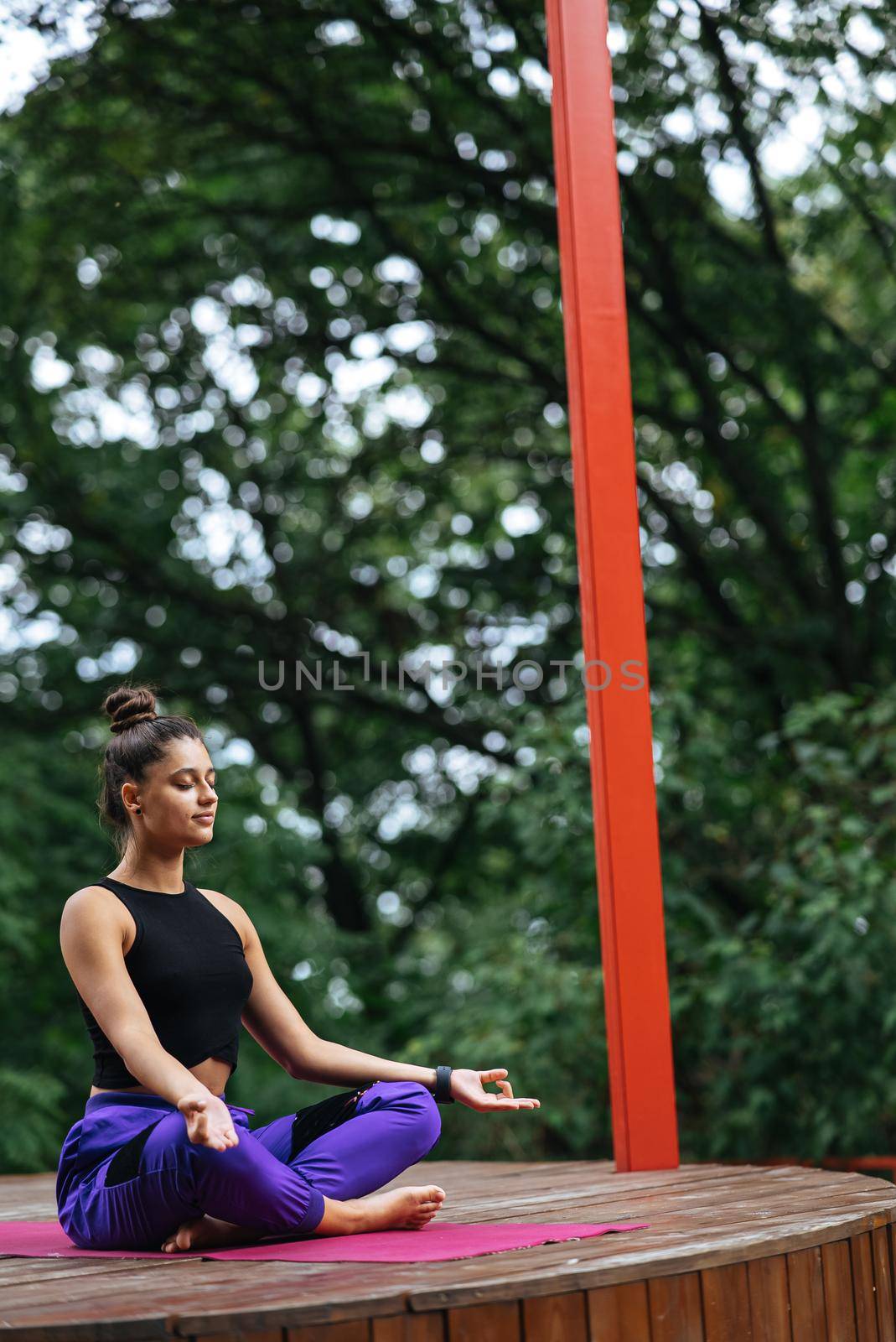 Young woman is practising yoga sitting in the lotus position on a wooden podium in the garden