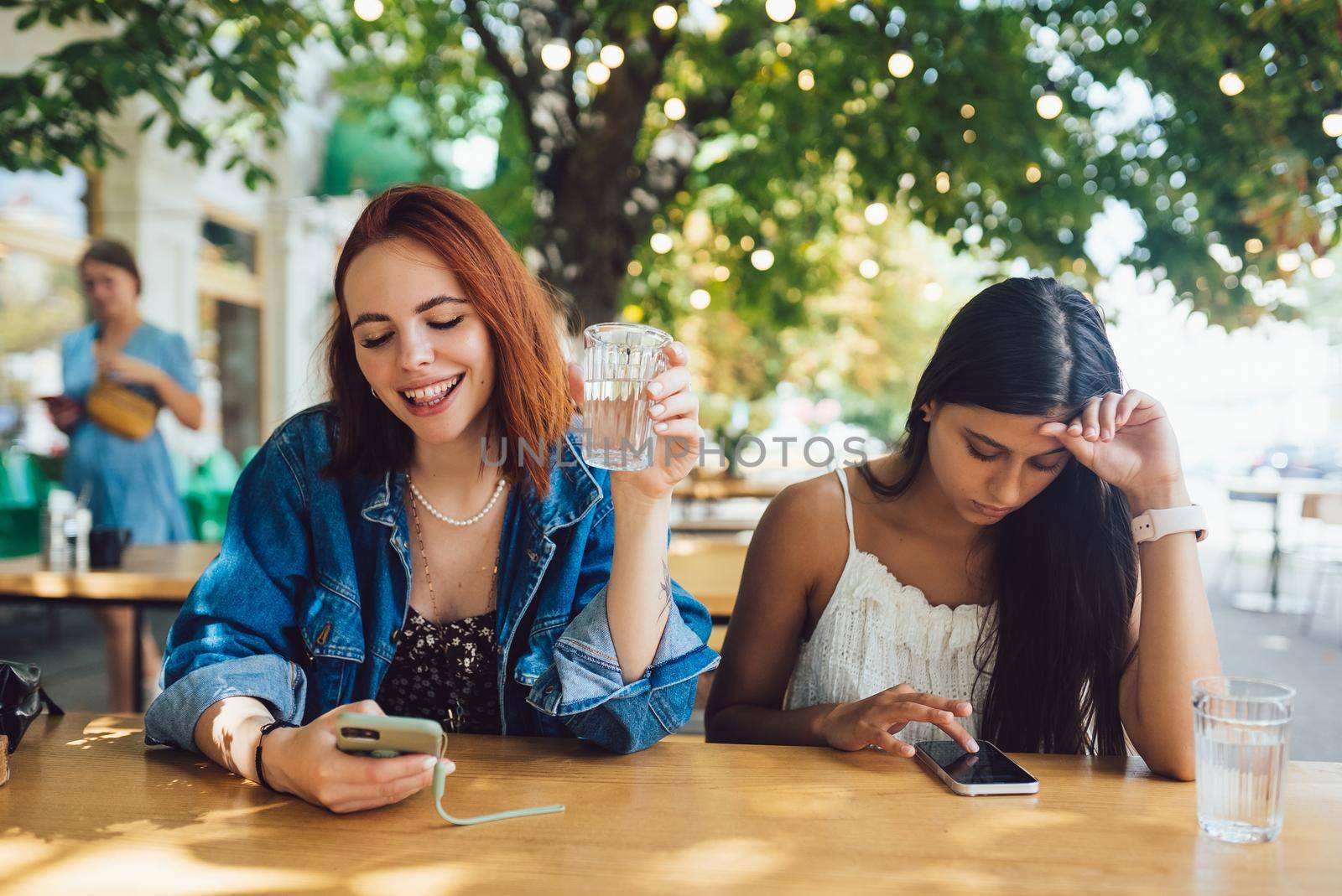 Two female friends browsing their mobile phones while sitting together at city street cafe. Girlfriends ignoring each other while staring in gadgets.