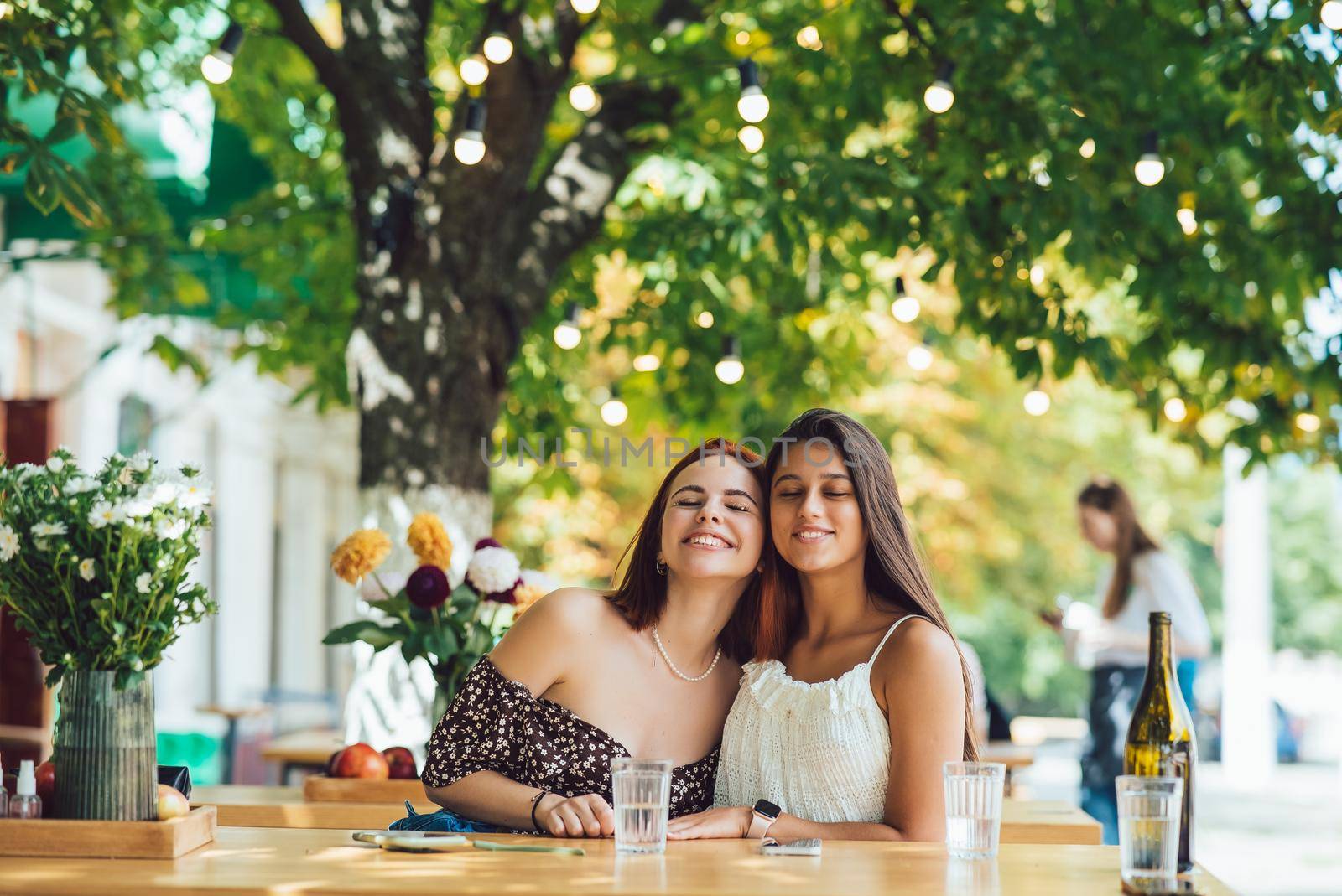 Close-up portrait of two female friends laughing drinking coffee on terrace at summer street cafe on background buildings