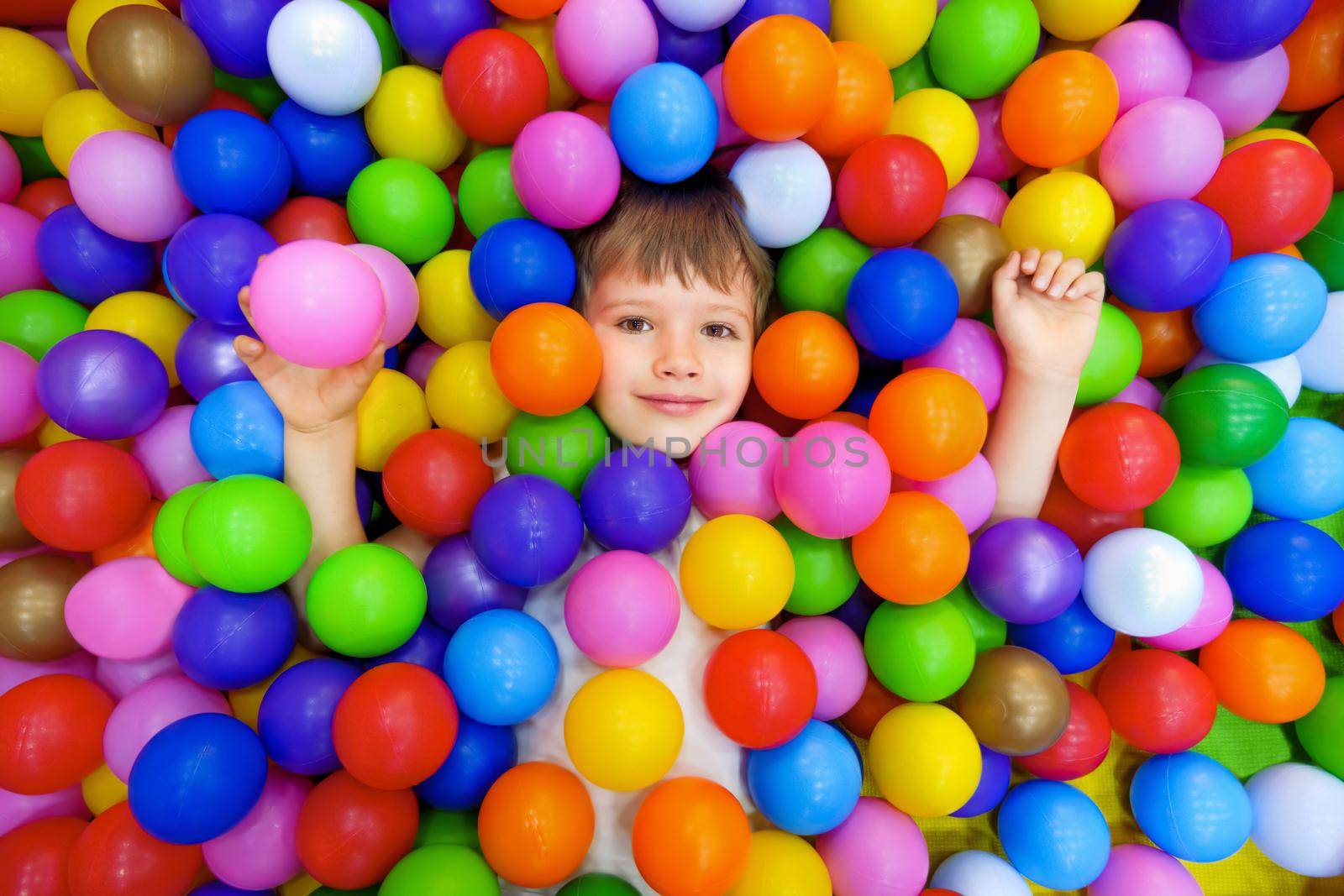 Smile kid lying colorful plastic balls pool. Colorful balls dry pool kindergarten playground child indoor play area. Playroom kids ball pit. Caucasian boy indoor playground kids play zone or kids zone