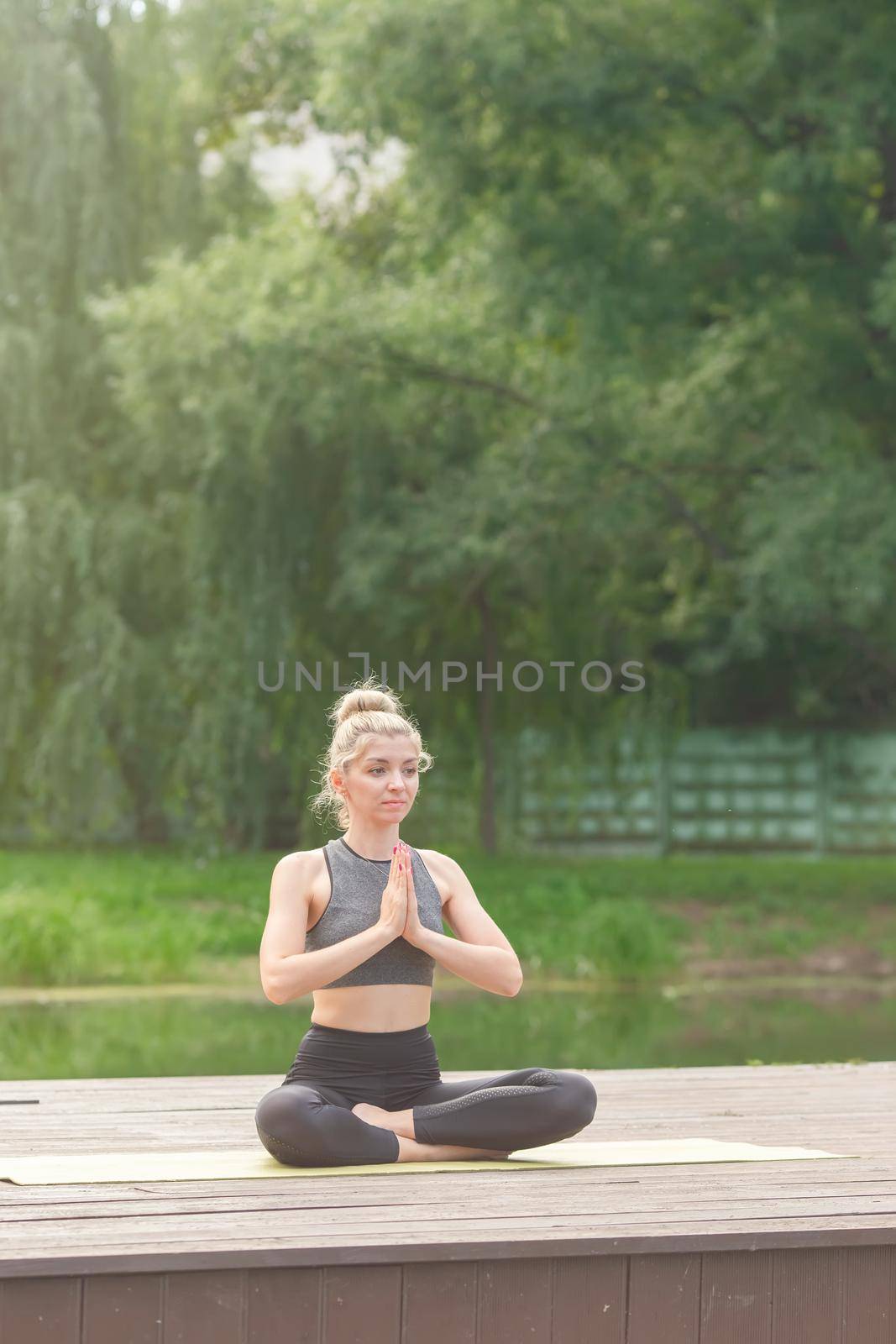 A beautiful woman in a gray top and leggings, sitting on a wooden platform by a pond in the park in summer, does yoga . Vertical. Copy space