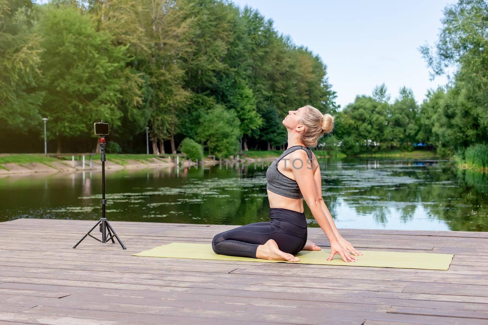 A slender woman in a gray top and leggings, sits on a wooden platform by a pond in the park in summer, does yoga, performs exercises, on a sports mat, in front of her smartphone on a tripod.