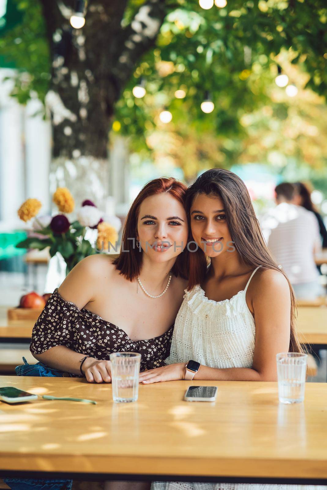 Close-up portrait of two female friends laughing drinking coffee on terrace at summer street cafe on background buildings