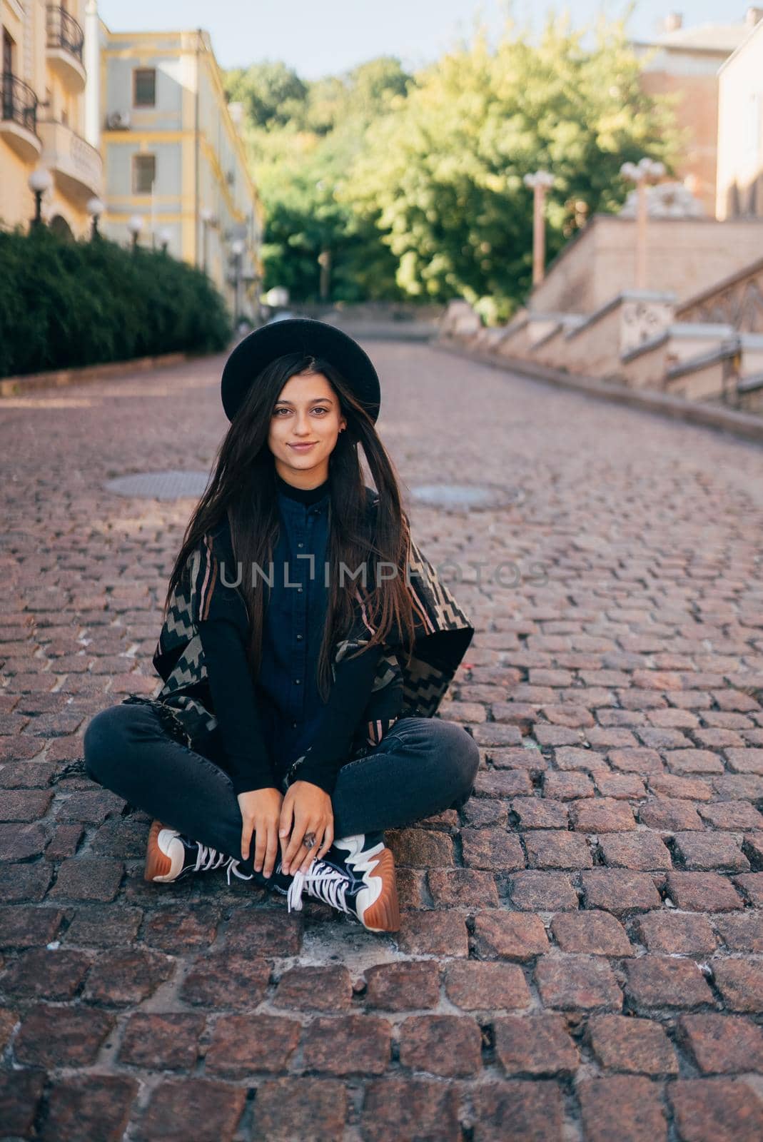 Young woman in hat sitting at city on cobblestone, warm autumn day