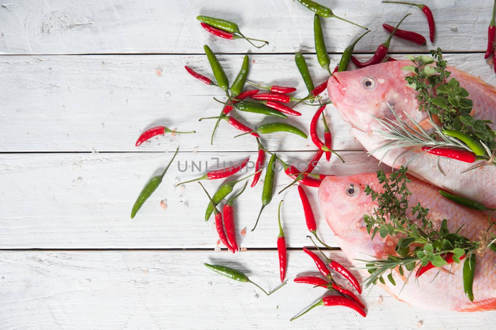 raw fresh pink tilapia fish lies on a board sprinkled with small red and green hot peppers, pink Himalayan salt and garnished with fresh curry sprigs, on white wooden planks. High quality photo