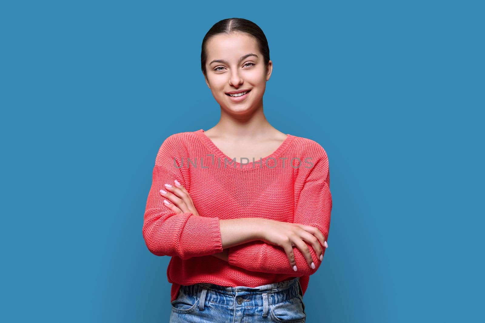 Portrait of teenage smiling female looking at camera on blue color studio background. Confident teen girl with crossed arms in red. Adolescence, high school, youth 15, 16 years old concept