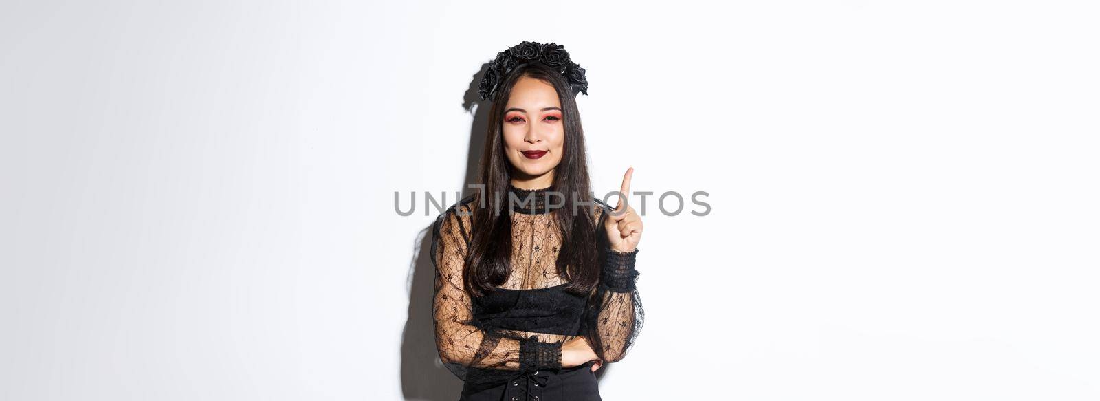 Creative young woman in witch costume smiling pleased as have great idea, raising finger to say suggestion. Female asian dressed-up like widow or mysterious magician, white background by Benzoix