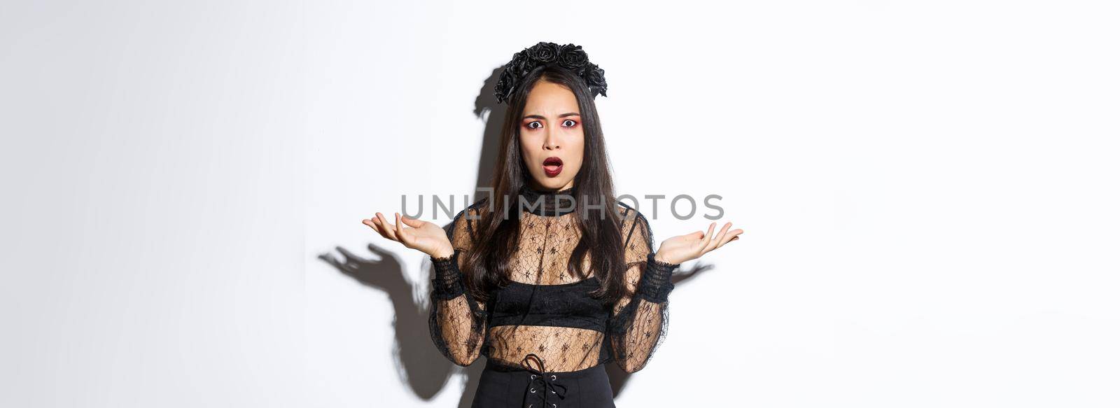 Confused asian woman in halloween costume looking clueless, raising hands sideways and stare puzzled at camera. Female wearing black lace dress, being a witch or magician on trick or treat party by Benzoix
