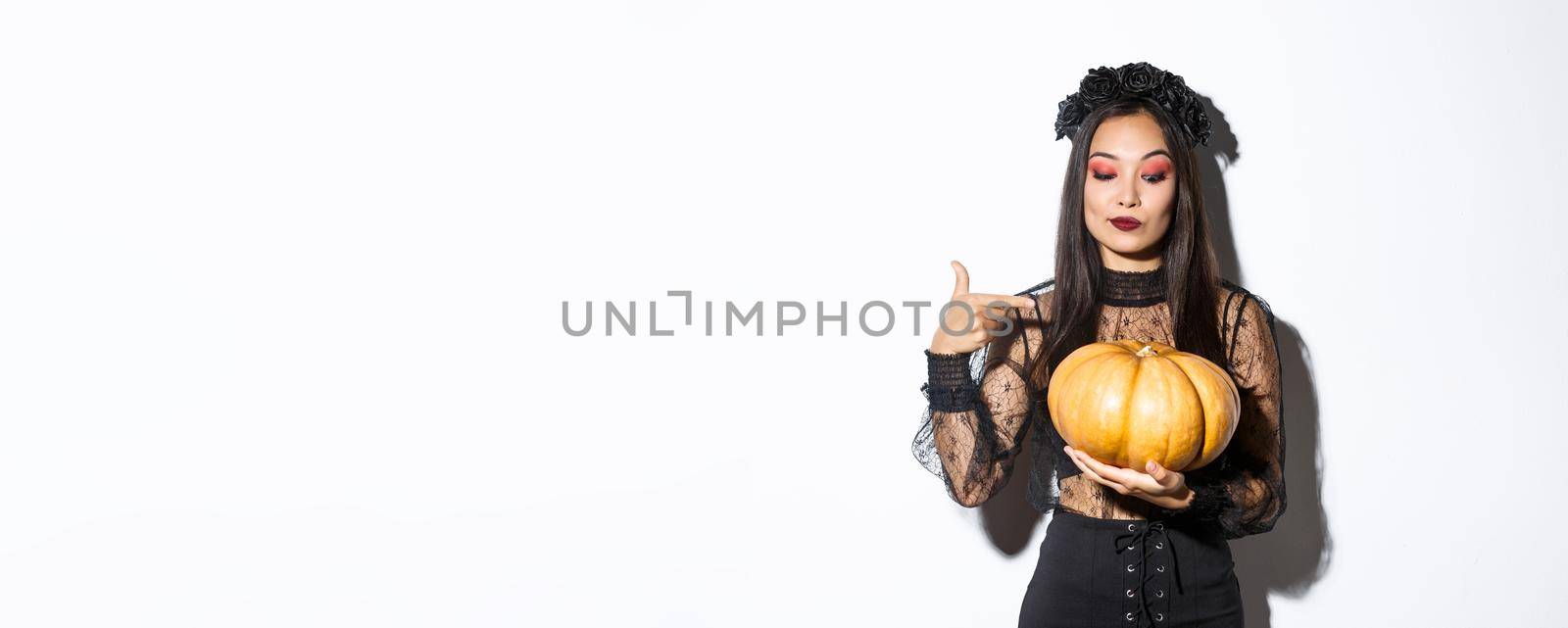 Image of beautiful asian woman in gothic lace dress and black wreath pointing finger at big pumpkin, celebrating halloween, standing over white background.