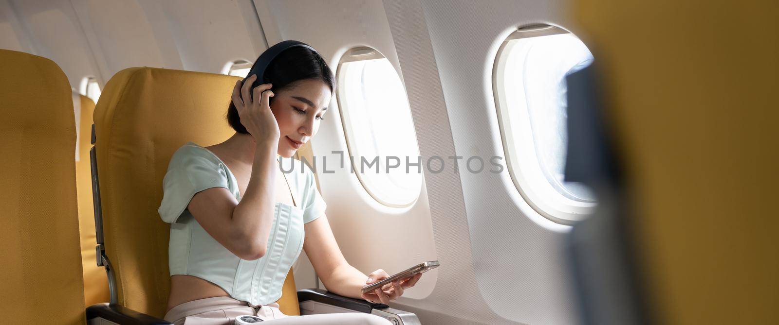 Young woman with mobile phone and headphones listening to music in airplane during flight by nateemee