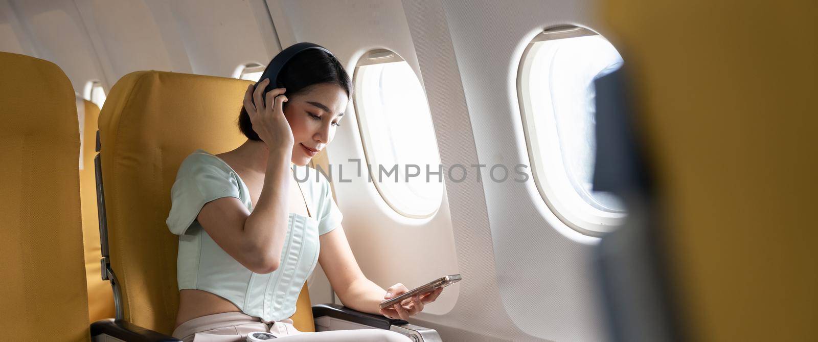 Young woman with mobile phone and headphones listening to music in airplane during flight.